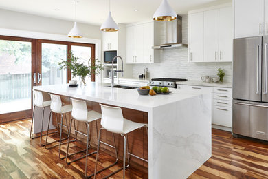 Photo of a transitional galley kitchen in Toronto with a double-bowl sink, flat-panel cabinets, white cabinets, white splashback, matchstick tile splashback, stainless steel appliances, medium hardwood floors, with island and brown floor.