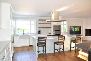 This is an example of a medium sized contemporary open plan kitchen in Other with a belfast sink, recessed-panel cabinets, white cabinets, quartz worktops, white splashback, engineered quartz splashback, stainless steel appliances, medium hardwood flooring, an island, brown floors and white worktops.