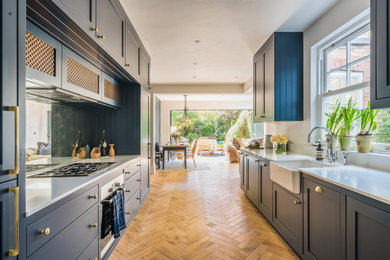 This is an example of a mid-sized traditional galley eat-in kitchen in London with a farmhouse sink, shaker cabinets, blue cabinets, solid surface benchtops, mirror splashback, stainless steel appliances, medium hardwood floors, no island, multi-coloured floor and white benchtop.