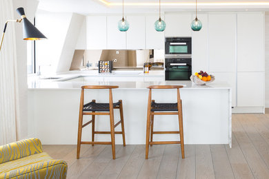 Design ideas for a large contemporary u-shaped open plan kitchen in London with a built-in sink, flat-panel cabinets, white cabinets, stainless steel appliances, light hardwood flooring and a breakfast bar.