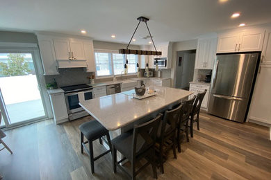 Photo of a transitional kitchen in Philadelphia with a farmhouse sink, flat-panel cabinets, white cabinets, quartz benchtops, grey splashback, subway tile splashback, stainless steel appliances, vinyl floors, with island, brown floor and grey benchtop.