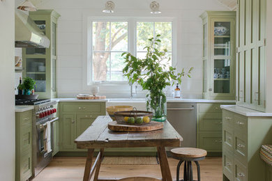 This is an example of a country kitchen in New York with a farmhouse sink, shaker cabinets, green cabinets, stainless steel appliances, light hardwood floors and no island.