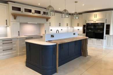 This is an example of an expansive traditional u-shaped open plan kitchen in Other with a belfast sink, shaker cabinets, beige cabinets, quartz worktops, white splashback, engineered quartz splashback, black appliances, an island, white worktops and feature lighting.