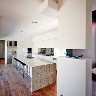 Modern inredning av ett stort beige linjärt beige skafferi, med en undermonterad diskho, luckor med infälld panel, vita skåp, bänkskiva i terrazo, stänkskydd med metallisk yta, glaspanel som stänkskydd, rostfria vitvaror, målat trägolv, en köksö och brunt golv