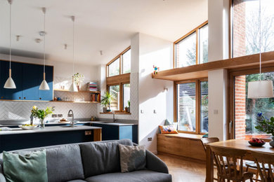 Photo of an expansive contemporary l-shaped open plan kitchen in London with an undermount sink, flat-panel cabinets, blue cabinets, quartzite benchtops, white splashback, ceramic splashback, black appliances, medium hardwood floors, with island, brown floor, white benchtop and vaulted.