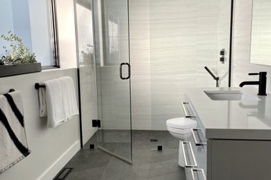 Cette image montre une salle de bain principale minimaliste de taille moyenne avec un placard à porte plane, des portes de placard grises, WC à poser, un carrelage blanc, un mur blanc, un sol en carrelage de porcelaine, un lavabo encastré, un plan de toilette en quartz modifié, un sol gris, une cabine de douche à porte battante, un plan de toilette blanc, une niche, meuble simple vasque et meuble-lavabo suspendu.