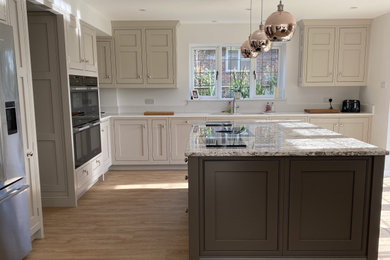 This is an example of an expansive traditional grey and brown u-shaped kitchen in Sussex with a double-bowl sink, shaker cabinets, beige cabinets, white splashback, tonge and groove splashback, black appliances, lino flooring, an island, brown floors and white worktops.