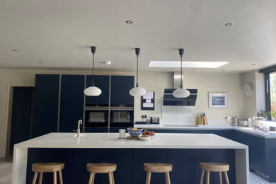 Large modern l-shaped open plan kitchen in Sussex with a submerged sink, flat-panel cabinets, blue cabinets, quartz worktops, white splashback, integrated appliances, porcelain flooring, an island, grey floors, white worktops and feature lighting.