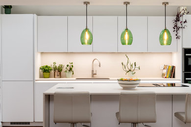 Large contemporary kitchen/diner in Kent with a single-bowl sink, flat-panel cabinets, white cabinets, composite countertops, white splashback, glass sheet splashback, integrated appliances, medium hardwood flooring, an island, brown floors and beige worktops.