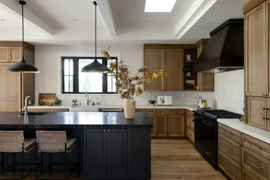 Large transitional l-shaped medium tone wood floor, brown floor and tray ceiling open concept kitchen photo in San Francisco with a farmhouse sink, shaker cabinets, medium tone wood cabinets, quartzite countertops, white backsplash, ceramic backsplash, black appliances, an island and black countertops