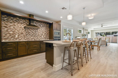 This is an example of a classic l-shaped open plan kitchen in Orlando with a double-bowl sink, shaker cabinets, medium wood cabinets, engineered stone countertops, metallic splashback, glass tiled splashback, stainless steel appliances, an island and grey floors.