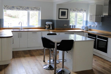 Design ideas for a large contemporary u-shaped open plan kitchen in Other with a double-bowl sink, white cabinets, wood worktops, blue splashback, glass sheet splashback, stainless steel appliances, medium hardwood flooring, an island, brown floors and flat-panel cabinets.