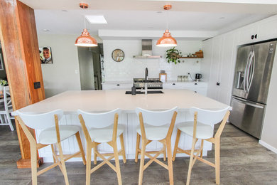 Photo of a large country l-shaped open plan kitchen in Sydney with a drop-in sink, shaker cabinets, white cabinets, quartz benchtops, white splashback, ceramic splashback, stainless steel appliances, light hardwood floors, with island, grey floor and white benchtop.