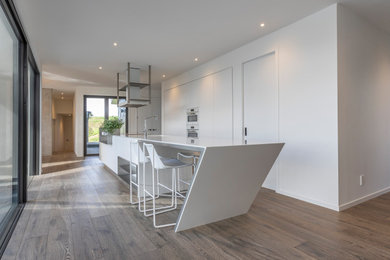 Large modern galley eat-in kitchen in Auckland with an undermount sink, flat-panel cabinets, yellow cabinets, solid surface benchtops, white appliances, medium hardwood floors, with island, brown floor and white benchtop.