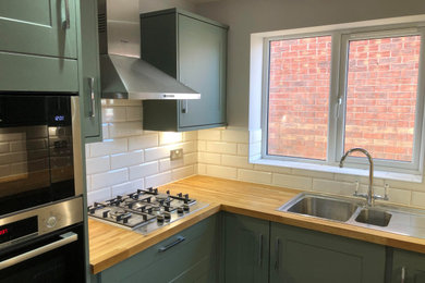 Photo of a small contemporary u-shaped open plan kitchen in Other with a double-bowl sink, shaker cabinets, green cabinets, wood worktops, yellow splashback, ceramic splashback, black appliances, laminate floors, no island, brown floors, brown worktops and feature lighting.