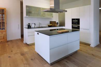 Mid-sized contemporary l-shaped open plan kitchen in Bremen with white cabinets, white splashback, black appliances, with island, a drop-in sink, light hardwood floors, brown floor, flat-panel cabinets and quartz benchtops.