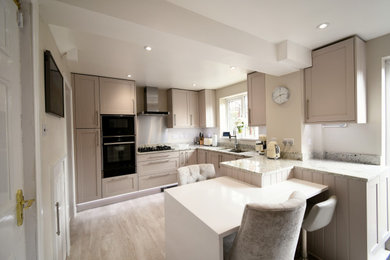 This is an example of a medium sized traditional l-shaped enclosed kitchen in Essex with a double-bowl sink, shaker cabinets, all types of cabinet finish, granite worktops, grey splashback, metal splashback, black appliances, laminate floors, a breakfast bar, white worktops and feature lighting.