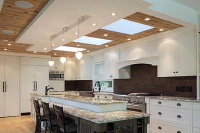Example of a mid-sized transitional galley light wood floor enclosed kitchen design in Other with a farmhouse sink, shaker cabinets, white cabinets, granite countertops, brown backsplash, stone tile backsplash, stainless steel appliances and an island