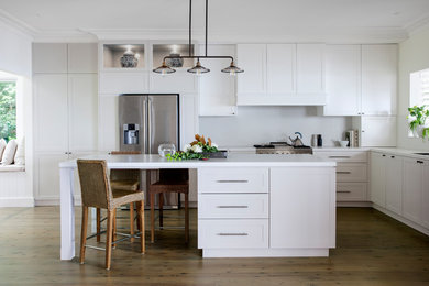 Design ideas for a transitional l-shaped kitchen in Sydney with an undermount sink, shaker cabinets, white cabinets, stainless steel appliances, medium hardwood floors, with island, brown floor and white benchtop.