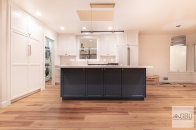 Mid-sized minimalist galley light wood floor and brown floor eat-in kitchen photo in San Francisco with shaker cabinets, white cabinets, quartz countertops, white backsplash, ceramic backsplash, white appliances, an island, white countertops and a farmhouse sink