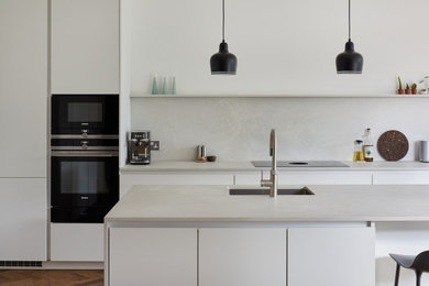 Mid-sized modern l-shaped open plan kitchen in London with flat-panel cabinets, white cabinets, quartz benchtops, grey splashback, with island and grey benchtop.