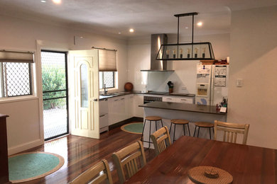 This is an example of a mid-sized contemporary l-shaped eat-in kitchen in Brisbane with a double-bowl sink, flat-panel cabinets, white cabinets, white splashback, glass sheet splashback, white appliances, medium hardwood floors and a peninsula.