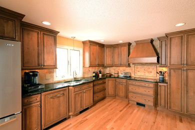 Large elegant l-shaped light wood floor open concept kitchen photo in Boston with an undermount sink, raised-panel cabinets, medium tone wood cabinets, granite countertops, beige backsplash, ceramic backsplash and stainless steel appliances