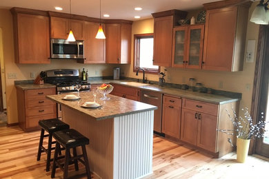 This is an example of a mid-sized transitional l-shaped eat-in kitchen in Milwaukee with an integrated sink, recessed-panel cabinets, medium wood cabinets, solid surface benchtops, stainless steel appliances, light hardwood floors, with island and brown floor.