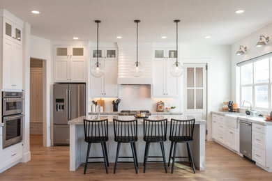 This is an example of a large transitional u-shaped separate kitchen in Salt Lake City with a farmhouse sink, shaker cabinets, white cabinets, marble benchtops, white splashback, stainless steel appliances, light hardwood floors, with island, beige floor, grey benchtop and porcelain splashback.