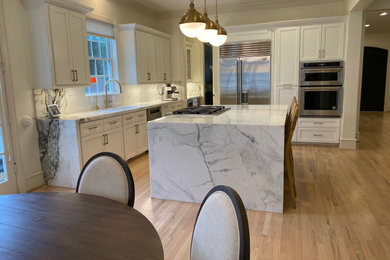 This is an example of a large contemporary l-shaped eat-in kitchen in Houston with a single-bowl sink, shaker cabinets, white cabinets, quartzite benchtops, white splashback, stainless steel appliances, light hardwood floors, with island and white benchtop.