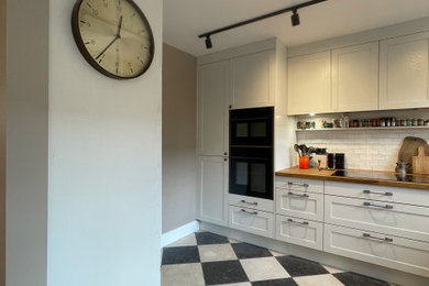 Photo of a medium sized traditional cream and black l-shaped kitchen/diner in London with a belfast sink, shaker cabinets, beige cabinets, wood worktops, white splashback, ceramic splashback, integrated appliances, limestone flooring, no island, black floors, brown worktops and feature lighting.