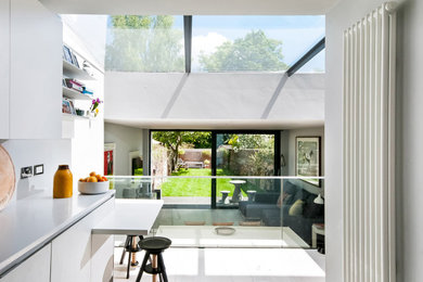 Medium sized contemporary grey and white single-wall open plan kitchen in Hampshire with an integrated sink, flat-panel cabinets, white cabinets, composite countertops, white splashback, integrated appliances, light hardwood flooring, an island and white worktops.
