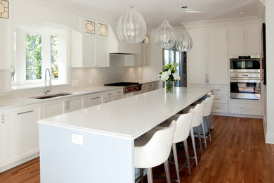 Photo of a mid-sized contemporary u-shaped open plan kitchen in Vancouver with a double-bowl sink, recessed-panel cabinets, white cabinets, solid surface benchtops, white splashback, ceramic splashback, stainless steel appliances, medium hardwood floors, with island, brown floor and white benchtop.