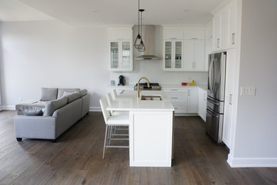 Design ideas for a mid-sized contemporary l-shaped open plan kitchen in Ottawa with an undermount sink, shaker cabinets, white cabinets, quartz benchtops, white splashback, ceramic splashback, stainless steel appliances, medium hardwood floors, with island, brown floor and white benchtop.