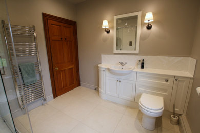 This is an example of a large classic ensuite bathroom in Oxfordshire with beaded cabinets, white cabinets, white tiles, solid surface worktops, white worktops, a single sink, a built in vanity unit, a corner shower, a one-piece toilet, ceramic tiles, grey walls, porcelain flooring, a built-in sink, beige floors and a sliding door.