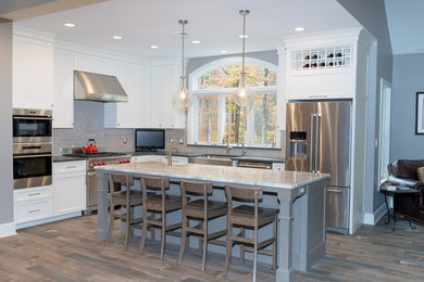 Example of a mid-sized transitional l-shaped brown floor open concept kitchen design in New York with a farmhouse sink, shaker cabinets, white cabinets, marble countertops, gray backsplash, ceramic backsplash, stainless steel appliances, an island and white countertops