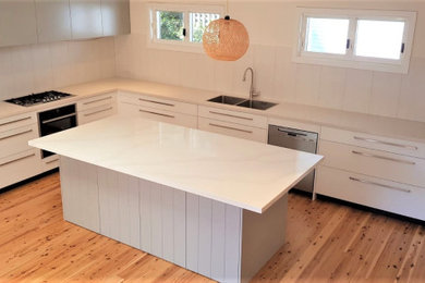 Design ideas for a mid-sized beach style l-shaped separate kitchen in Sydney with a double-bowl sink, flat-panel cabinets, white cabinets, marble benchtops, grey splashback, slate splashback, stainless steel appliances, medium hardwood floors, with island, beige floor and grey benchtop.