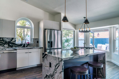Example of a mid-sized transitional l-shaped porcelain tile and brown floor eat-in kitchen design in Phoenix with an undermount sink, flat-panel cabinets, white cabinets, granite countertops, black backsplash, stone slab backsplash, stainless steel appliances, an island and black countertops