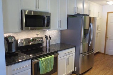 Inspiration for a mid-sized transitional medium tone wood floor and brown floor kitchen remodel in Milwaukee with an undermount sink, recessed-panel cabinets, white cabinets, granite countertops, white backsplash, glass tile backsplash, stainless steel appliances and black countertops