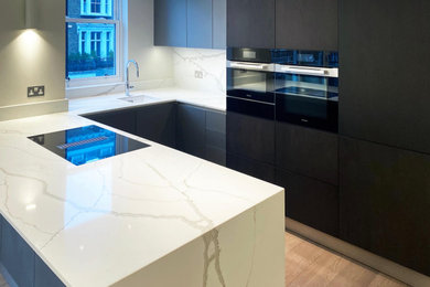 Large modern u-shaped open plan kitchen in London with a single-bowl sink, flat-panel cabinets, black cabinets, quartz worktops, white splashback, engineered quartz splashback, black appliances, light hardwood flooring, an island and white worktops.