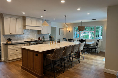 Example of a mid-sized transitional single-wall brown floor and medium tone wood floor eat-in kitchen design in Newark with shaker cabinets, white cabinets, quartz countertops, white backsplash, terra-cotta backsplash, stainless steel appliances, an island, black countertops and a farmhouse sink