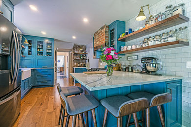 Mid-sized eclectic eat-in kitchen in Other with a farmhouse sink, shaker cabinets, blue cabinets, granite benchtops, white splashback, ceramic splashback, stainless steel appliances, medium hardwood floors, a peninsula, brown floor and grey benchtop.