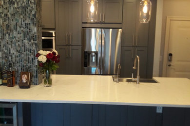 Small minimalist galley medium tone wood floor and gray floor eat-in kitchen photo in Atlanta with a farmhouse sink, shaker cabinets, gray cabinets, quartzite countertops, gray backsplash, glass sheet backsplash, stainless steel appliances and an island