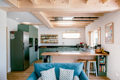 Photo of a contemporary open plan kitchen in West Midlands with a built-in sink, flat-panel cabinets, green cabinets, wood worktops, black splashback, granite splashback, stainless steel appliances, medium hardwood flooring, a breakfast bar, black worktops, exposed beams and feature lighting.
