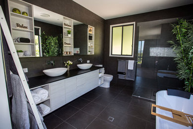 Mid-sized contemporary master bathroom in Sunshine Coast with furniture-like cabinets, grey cabinets, a freestanding tub, an open shower, a one-piece toilet, black tile, ceramic tile, black walls, ceramic floors, a vessel sink, engineered quartz benchtops, black floor, a hinged shower door and black benchtops.