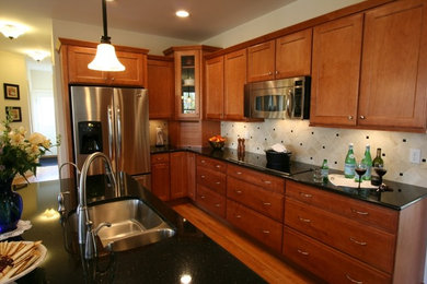 Photo of a large traditional l-shaped eat-in kitchen in New York with an undermount sink, granite benchtops, stone tile splashback, stainless steel appliances, shaker cabinets, medium wood cabinets, beige splashback, medium hardwood floors, with island, brown floor and black benchtop.