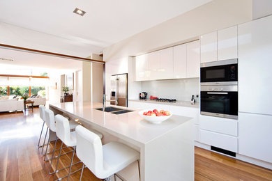 This is an example of a mid-sized modern single-wall open plan kitchen in Sydney with an undermount sink, beaded inset cabinets, white cabinets, quartzite benchtops, white splashback, glass sheet splashback, stainless steel appliances, medium hardwood floors and with island.