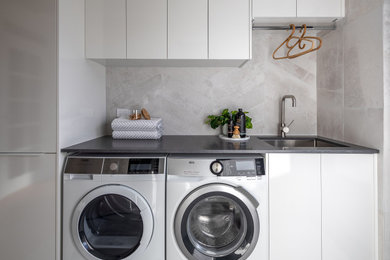 This is an example of a mid-sized modern single-wall laundry room in Perth with an undermount sink, white cabinets, grey splashback, ceramic splashback, grey walls, ceramic floors, a side-by-side washer and dryer, grey floor and grey benchtop.