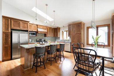 Example of a mid-sized transitional l-shaped medium tone wood floor, brown floor and vaulted ceiling eat-in kitchen design in Minneapolis with a double-bowl sink, shaker cabinets, medium tone wood cabinets, solid surface countertops, multicolored backsplash, ceramic backsplash, stainless steel appliances, an island and multicolored countertops