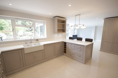Large contemporary u-shaped open plan kitchen in Surrey with a farmhouse sink, shaker cabinets, grey cabinets, solid surface benchtops, white splashback, stainless steel appliances, porcelain floors, a peninsula, beige floor and white benchtop.